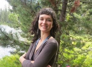 Megan with arms lightly crossed, smiling in front of a pine tree and lake.